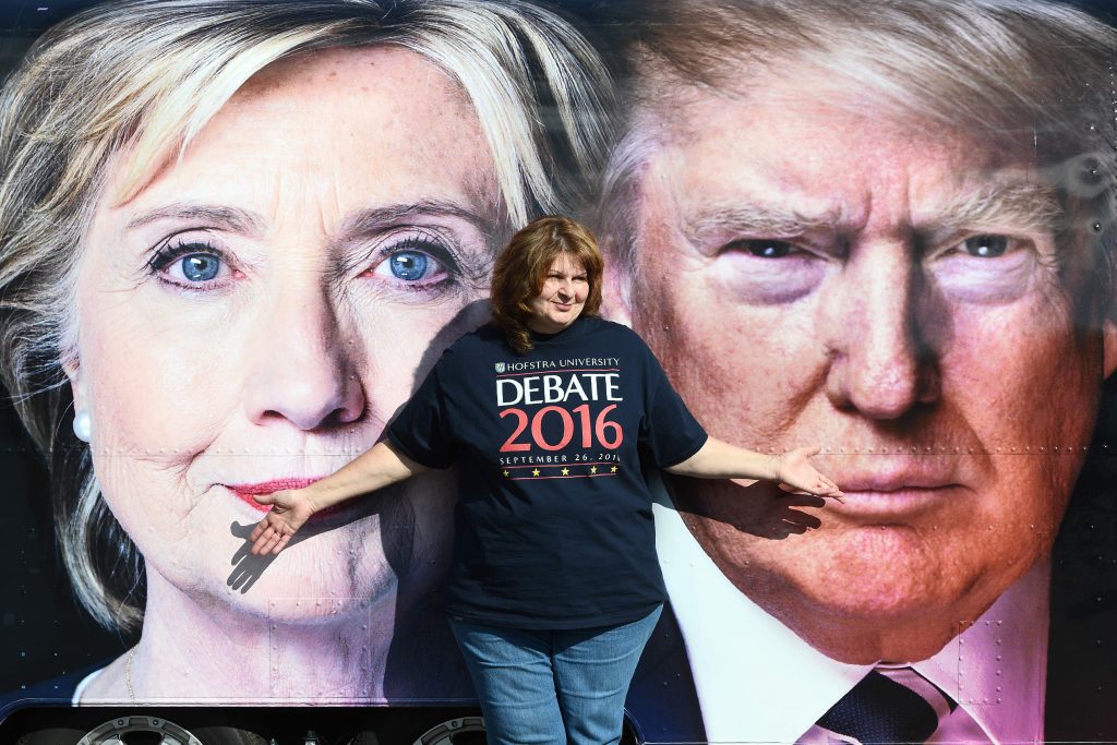 Mulher posa com a camiseta oficial diante de cartaz promovendo o debate entre Hillary e Trump